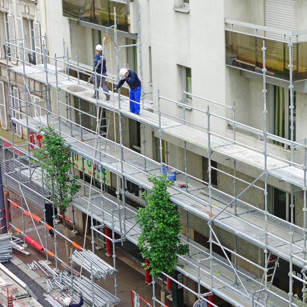 Chantier de rénovation urbaine: deux ouvriers sur un échafaudage métallique le long d'un bâtiment. Matériel de travaux au sol. Panneaux "CORDON NERVEUX".