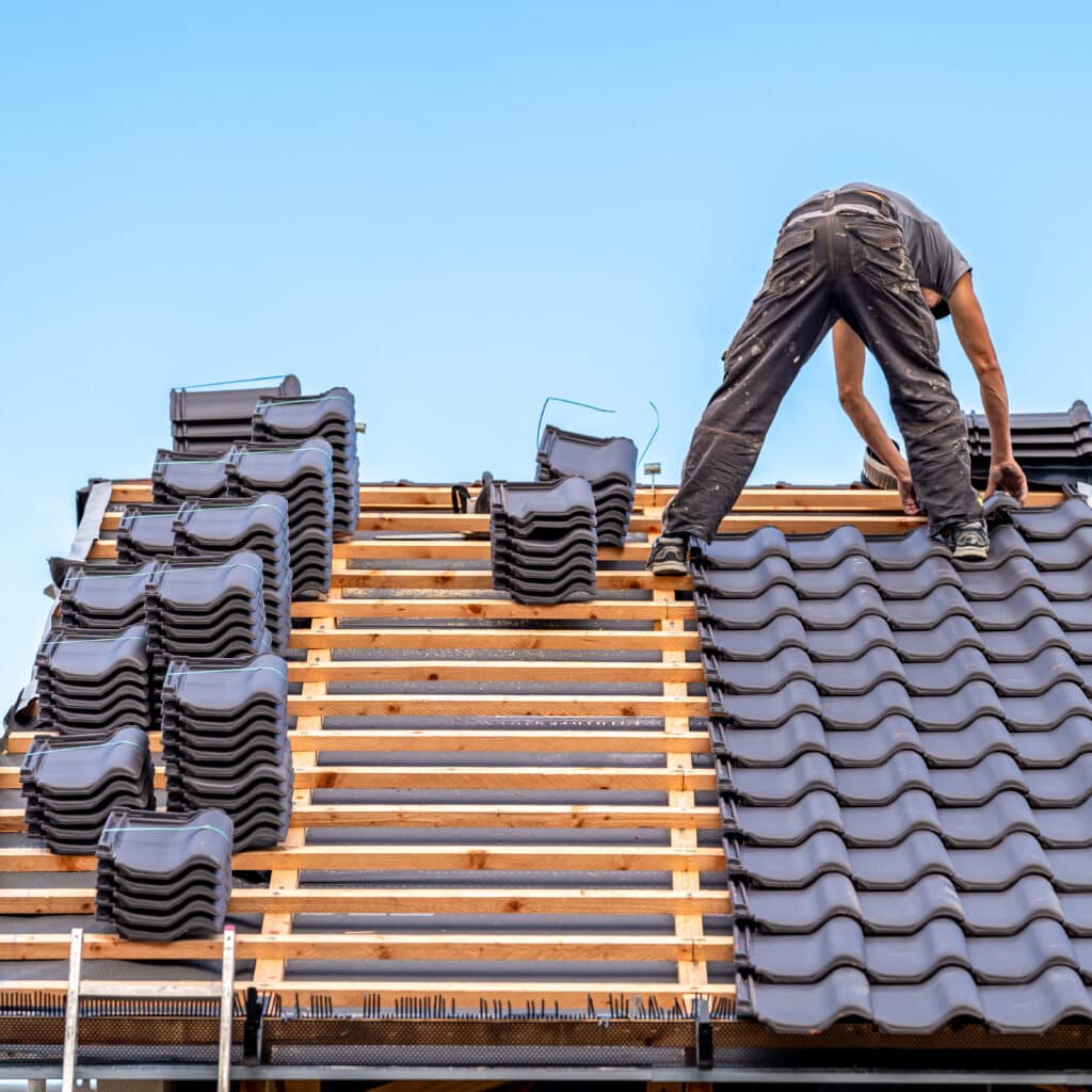 Couvreur installant des tuiles de toit grises sur une charpente en bois. Piles de tuiles attendent d'être posées sous un ciel bleu.