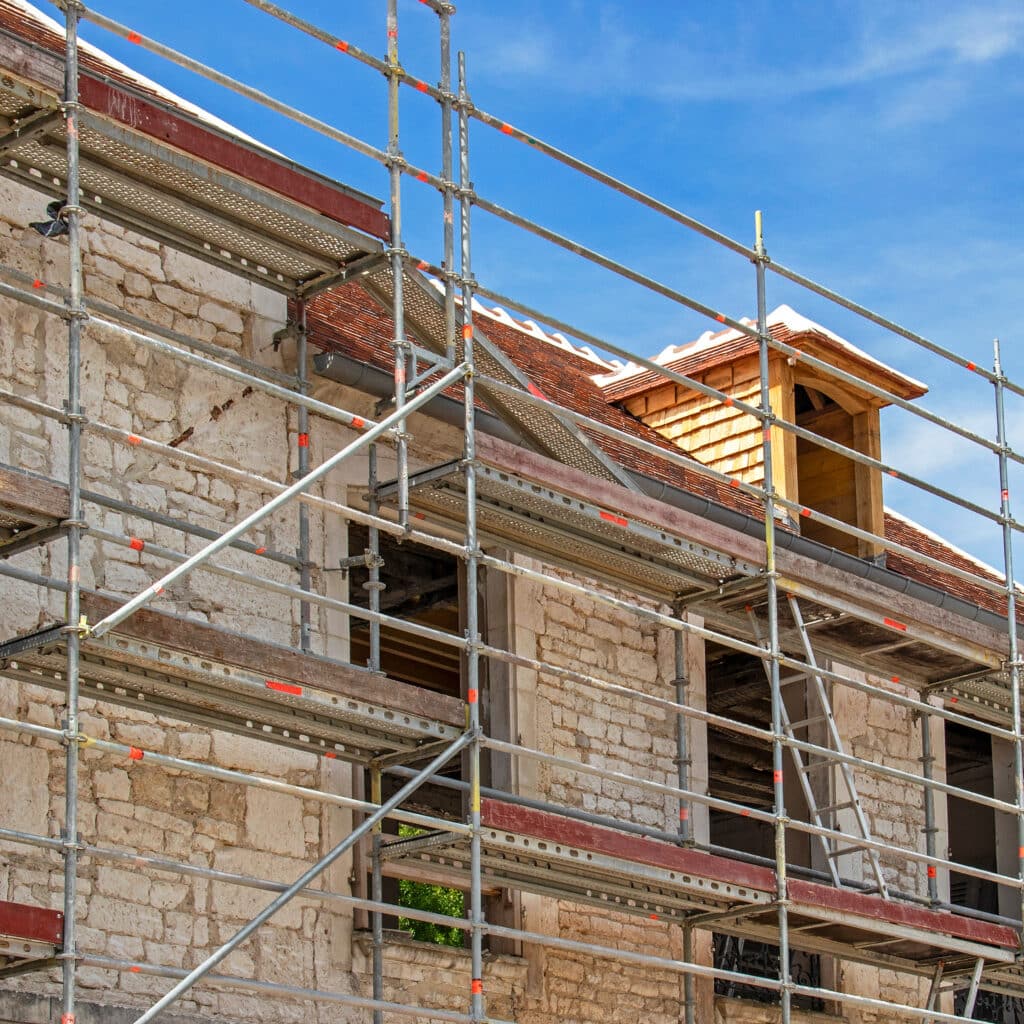 Échafaudage métallique sur un bâtiment en pierre en rénovation sous un ciel bleu. Toit en tuiles et lucarne en bois neufs.