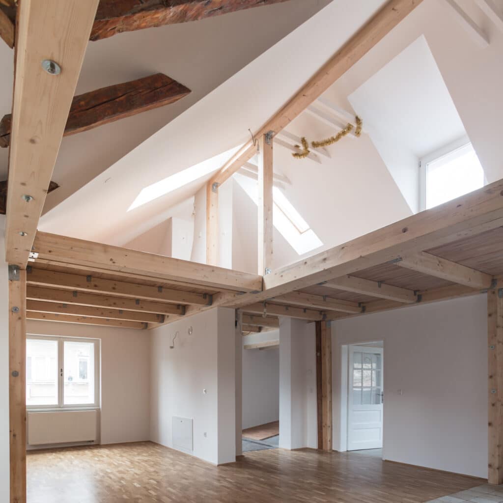 Aménagement de combles lumineux avec mezzanine en bois clair, poutres apparentes, murs blancs et parquet. Espace rénové.