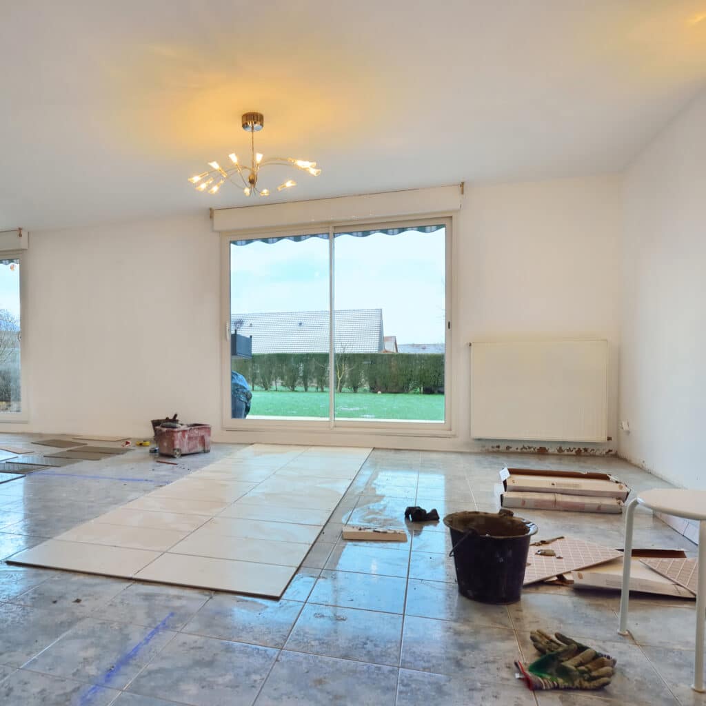 Pièce en rénovation: pose de carrelage clair sur sol ancien. Outils de chantier, seaux, gants de travail. Grandes baies vitrées avec vue jardin.
