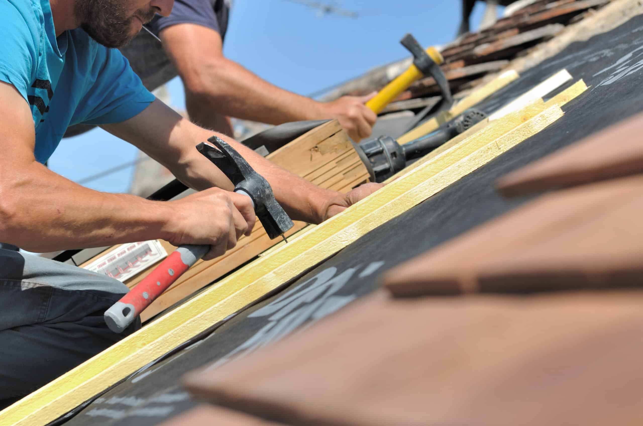 Un couvreur martèle une latte en bois sur un toit en rénovation avec membrane noire. Un second ouvrier est visible en arrière-plan sous un ciel bleu clair.