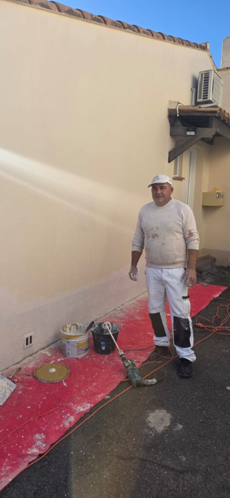 Peintre professionnel couvert d'enduit debout devant le mur texturé (numéro 6 visible), avec des seaux et un mélangeur sur une bâche rouge.